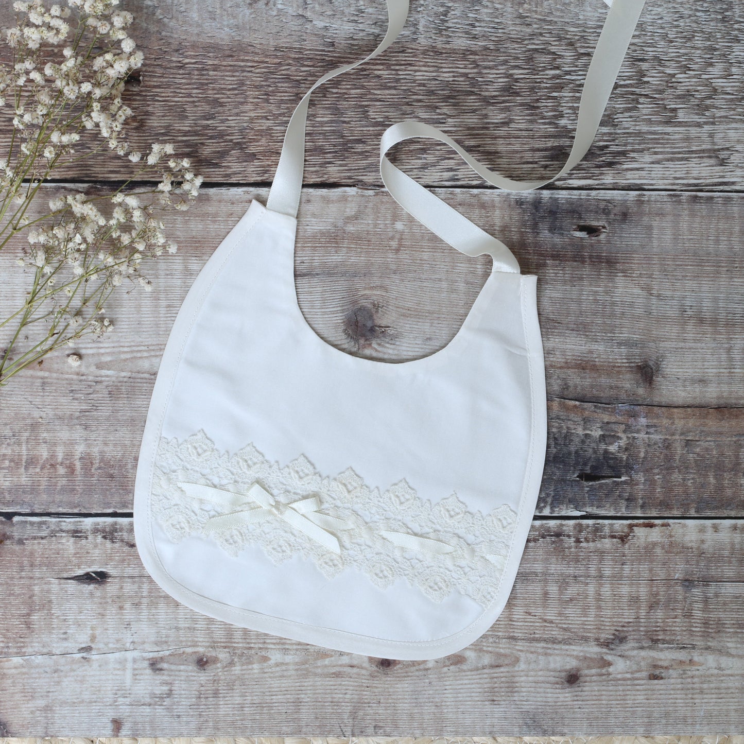 ivory baby bib with lace detailing on a wooden floor with flowers in the corner