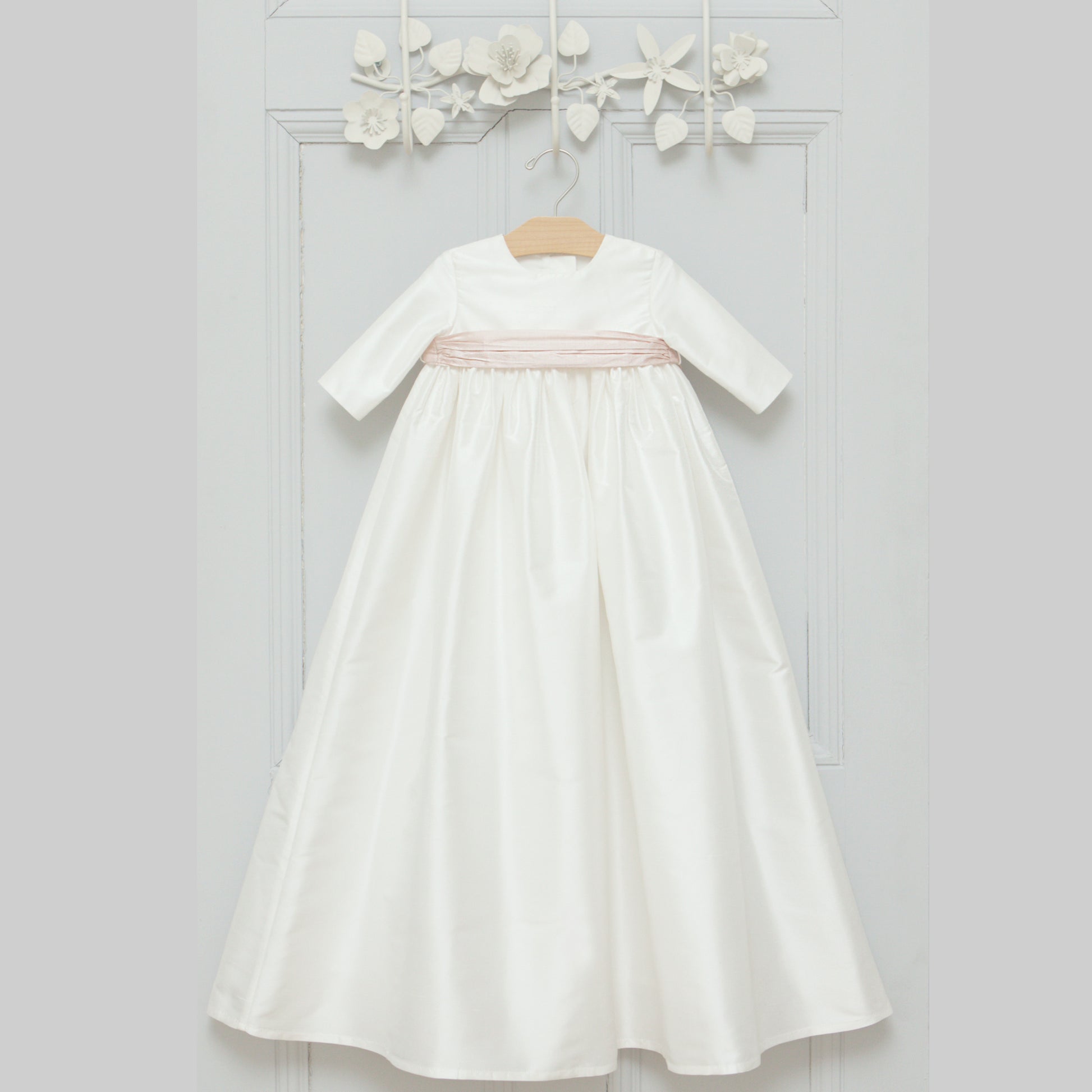 The Echo long sleeved christening gown with pink sash hanging on a grey door