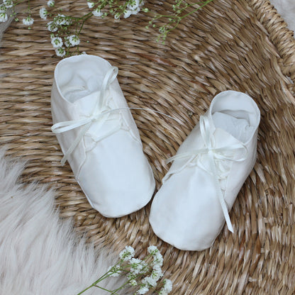 A pair of ivory Joshua booties with white satin ribbon laces, displayed on a round woven wicker tray with small white flowers and a white faux fur accent.