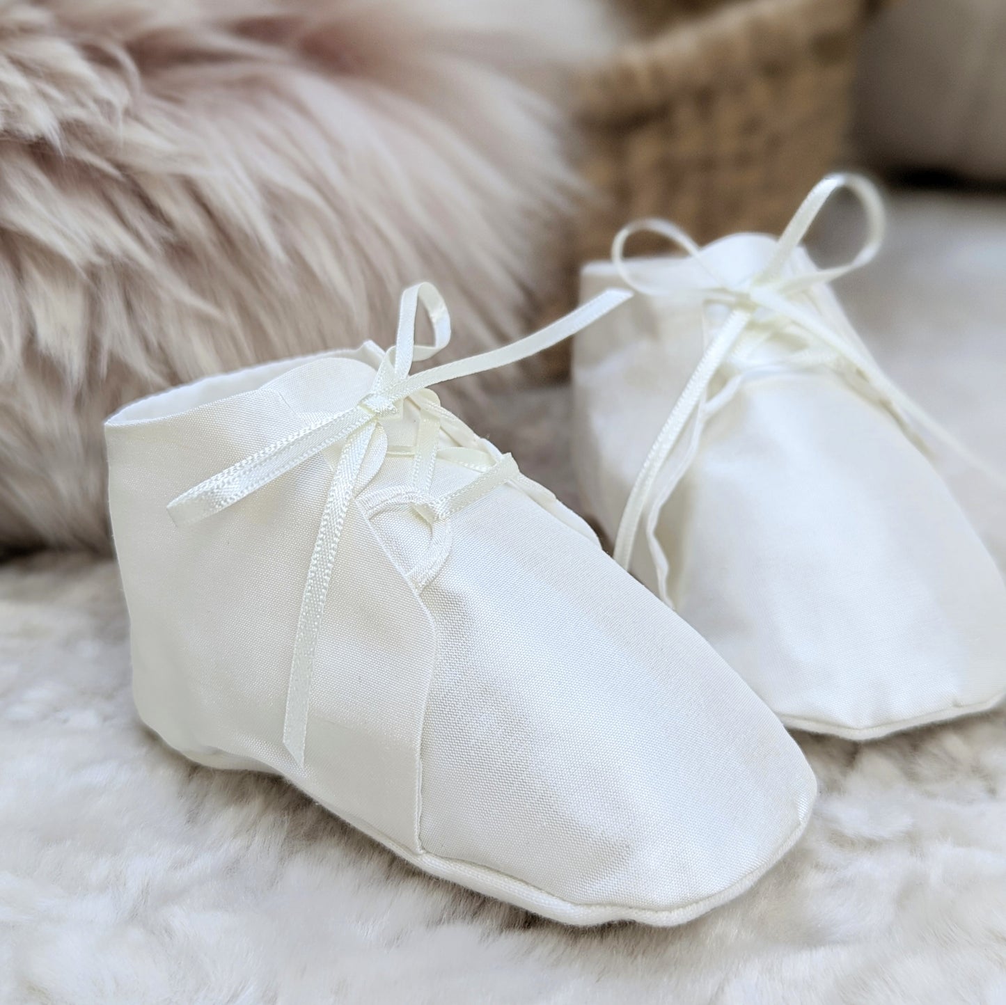 A pair of white fabric baby booties with ribbon laces, resting on a soft white faux fur surface with a neutral woven basket and fluffy beige background.
