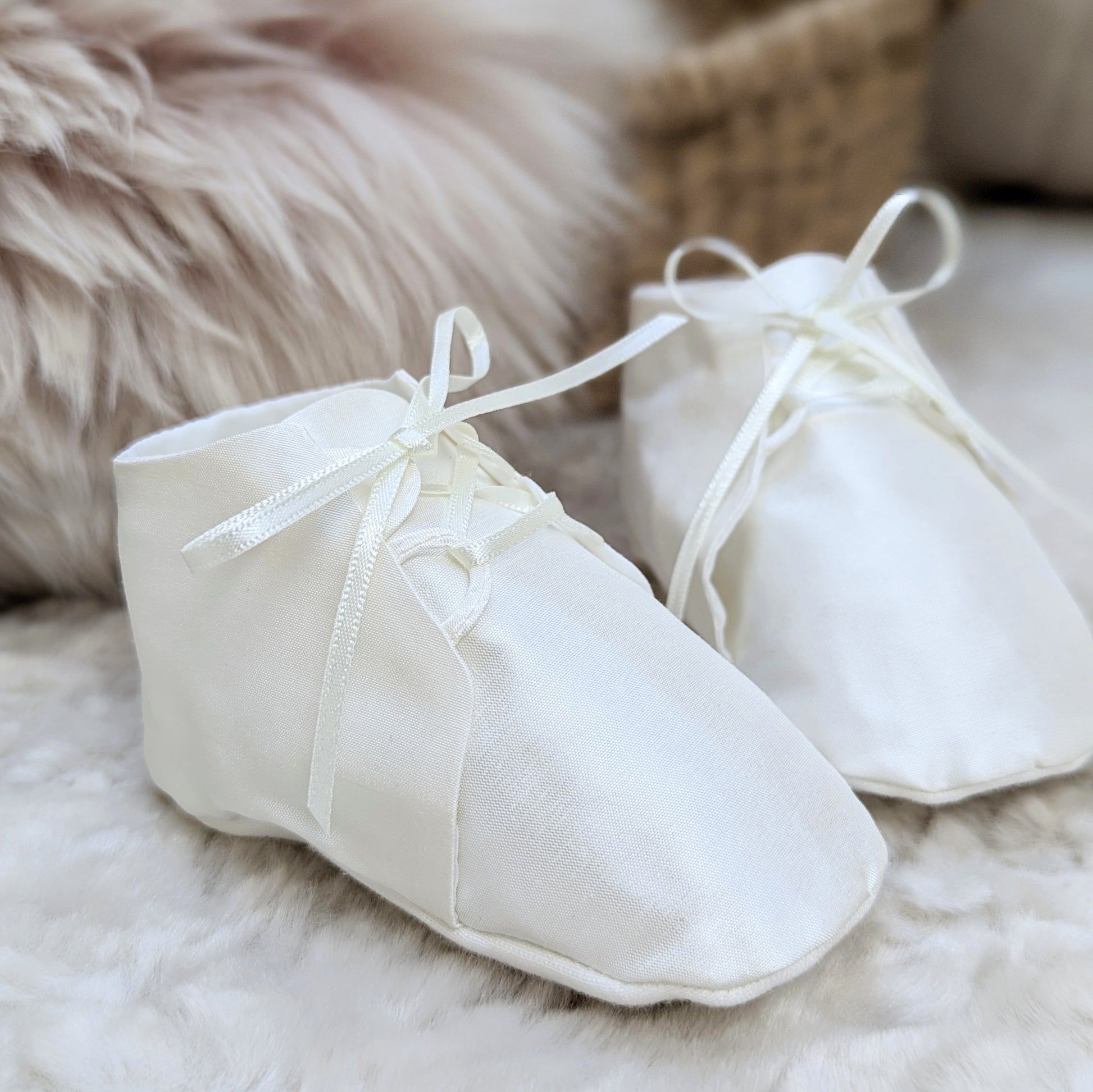 A pair of white fabric baby booties with ribbon laces, resting on a soft white faux fur surface with a neutral woven basket and fluffy beige background.