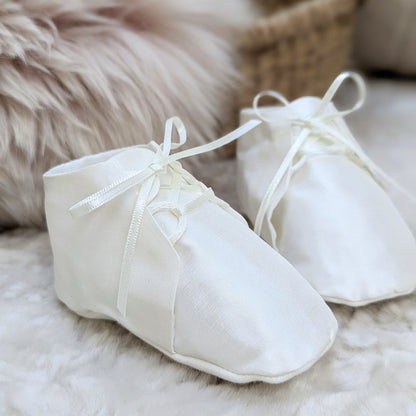 A pair of white fabric baby booties with ribbon laces, resting on a soft white faux fur surface with a neutral woven basket and fluffy beige background.