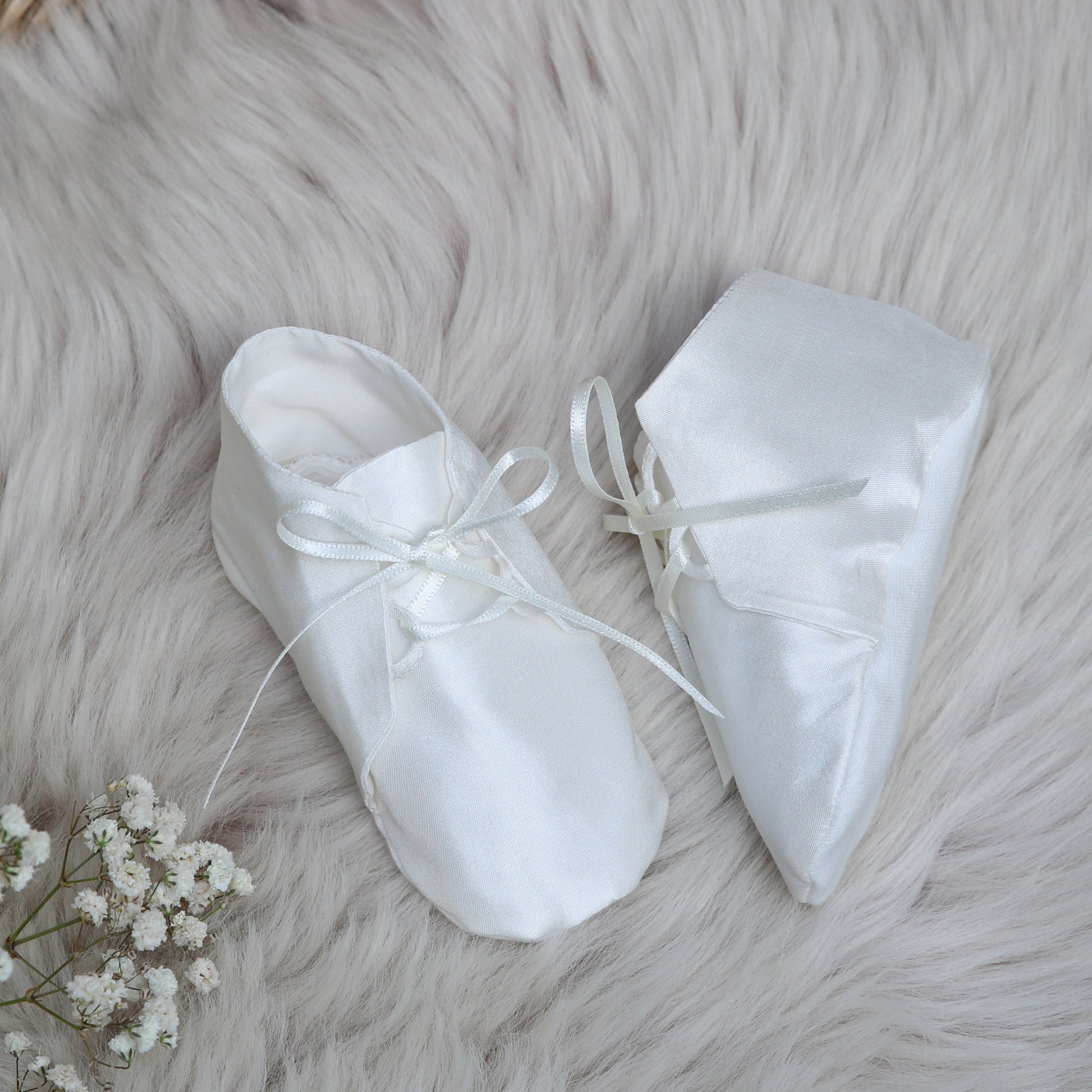A pair of Joshua ivory baby booties with ribbon laces, arranged on a fluffy white faux fur rug with baby's breath flowers and a woven mat in the background.