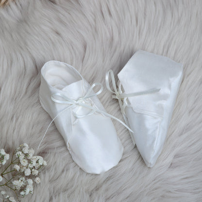 A pair of Joshua ivory baby booties with ribbon laces, arranged on a fluffy white faux fur rug with baby's breath flowers and a woven mat in the background.