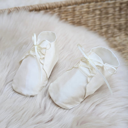 A pair of cream satin baby booties with matching ribbon laces, resting on a white faux fur rug next to a woven wicker basket.