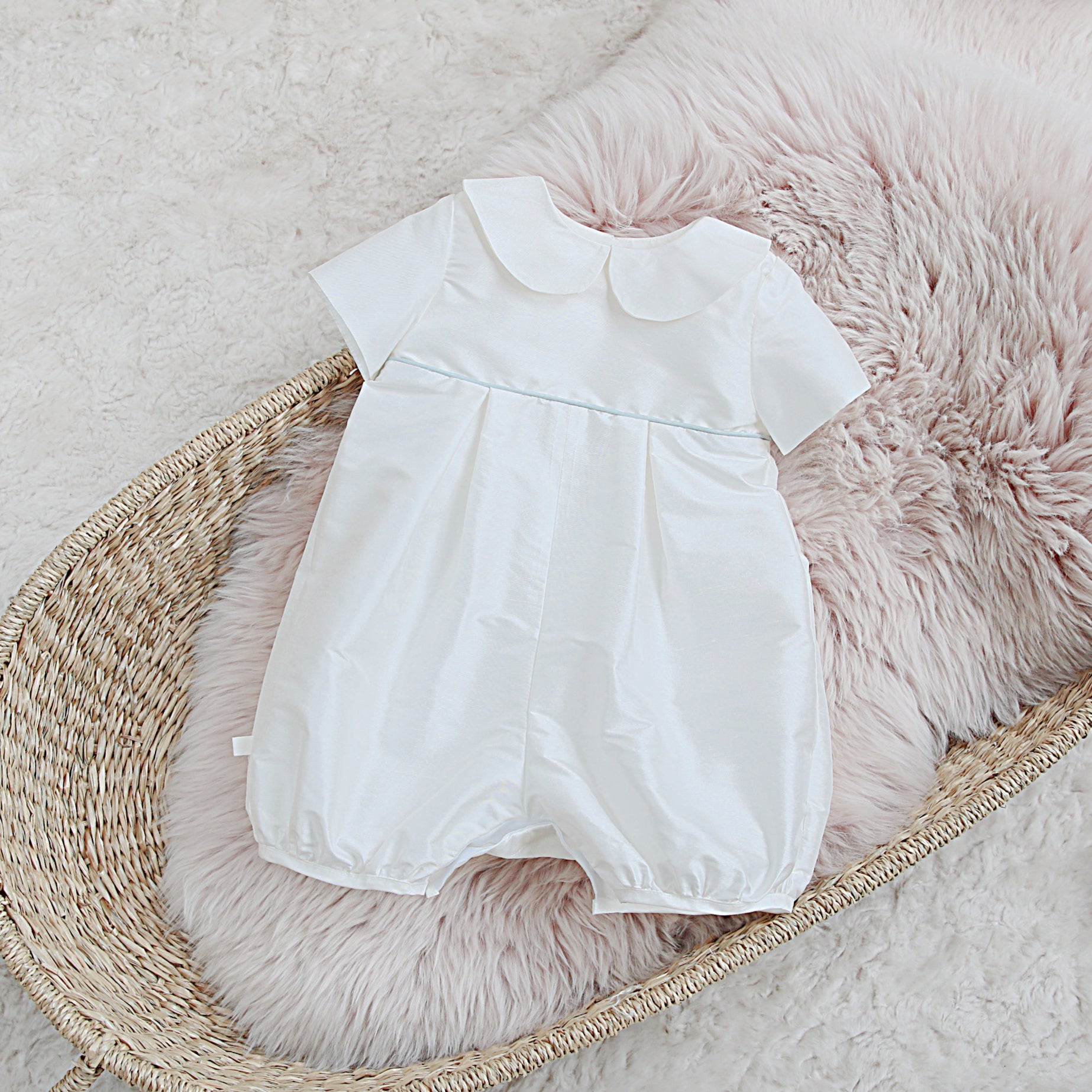 A white Joshua short-sleeve baby romper with a Peter Pan collar and a thin light blue horizontal stripe across the chest, displayed on a pink faux fur cushion inside a woven basket.