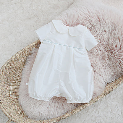 A white Joshua short-sleeve baby romper with a Peter Pan collar and a thin light blue horizontal stripe across the chest, displayed on a pink faux fur cushion inside a woven basket.