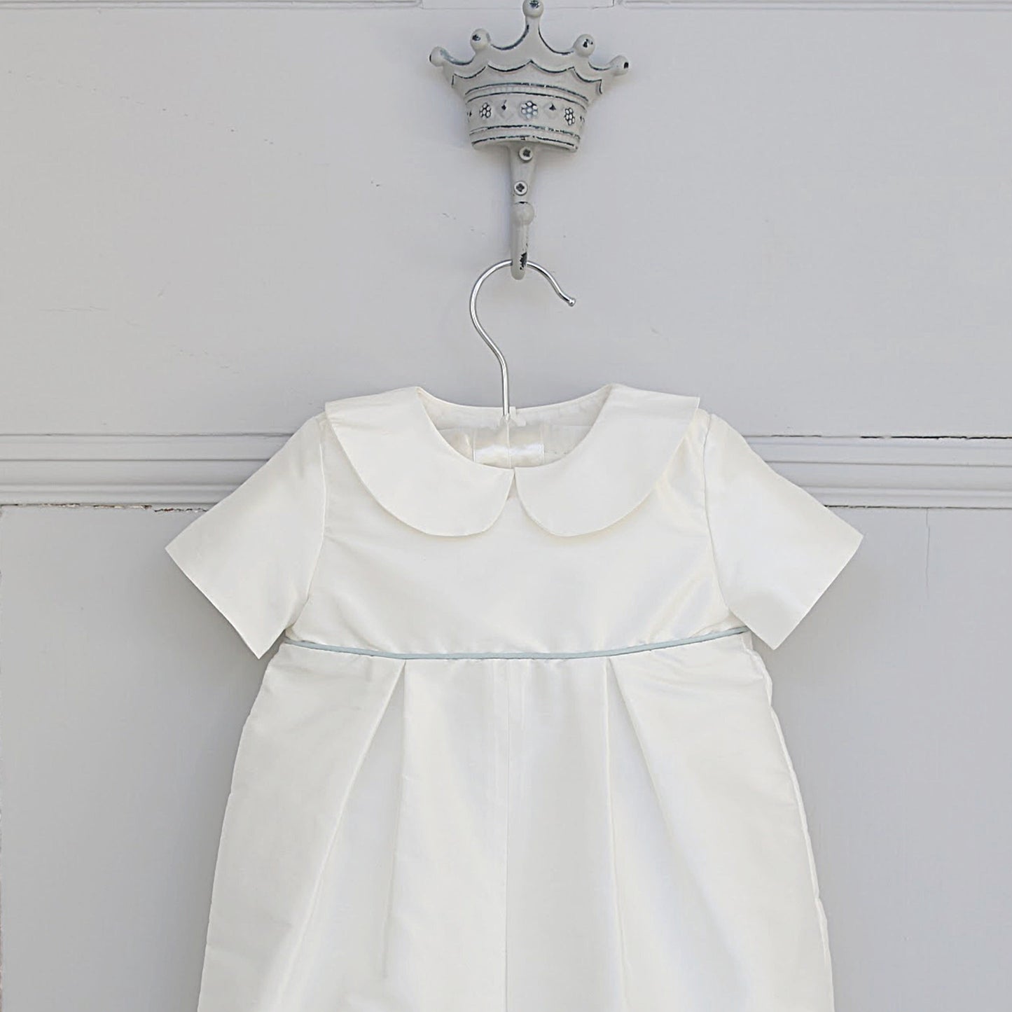 A white short-sleeve baby dress with a large Peter Pan collar and a thin light blue waistband, hanging on a silver hanger from a decorative crown-shaped wall hook against a white paneled wall.