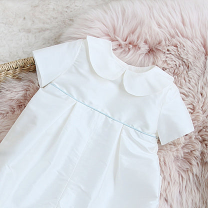 Joshua short-sleeve baby romper with a Peter Pan collar and a thin light blue piping detail, laid flat on a pink faux fur rug.