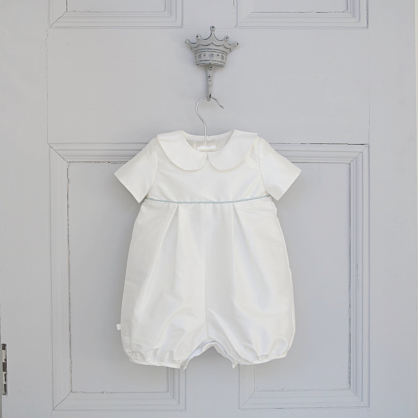 A white baby romper with short sleeves, a Peter Pan collar, and a subtle light blue piping detail at the waist, hanging on a decorative silver crown hook against a white paneled door.