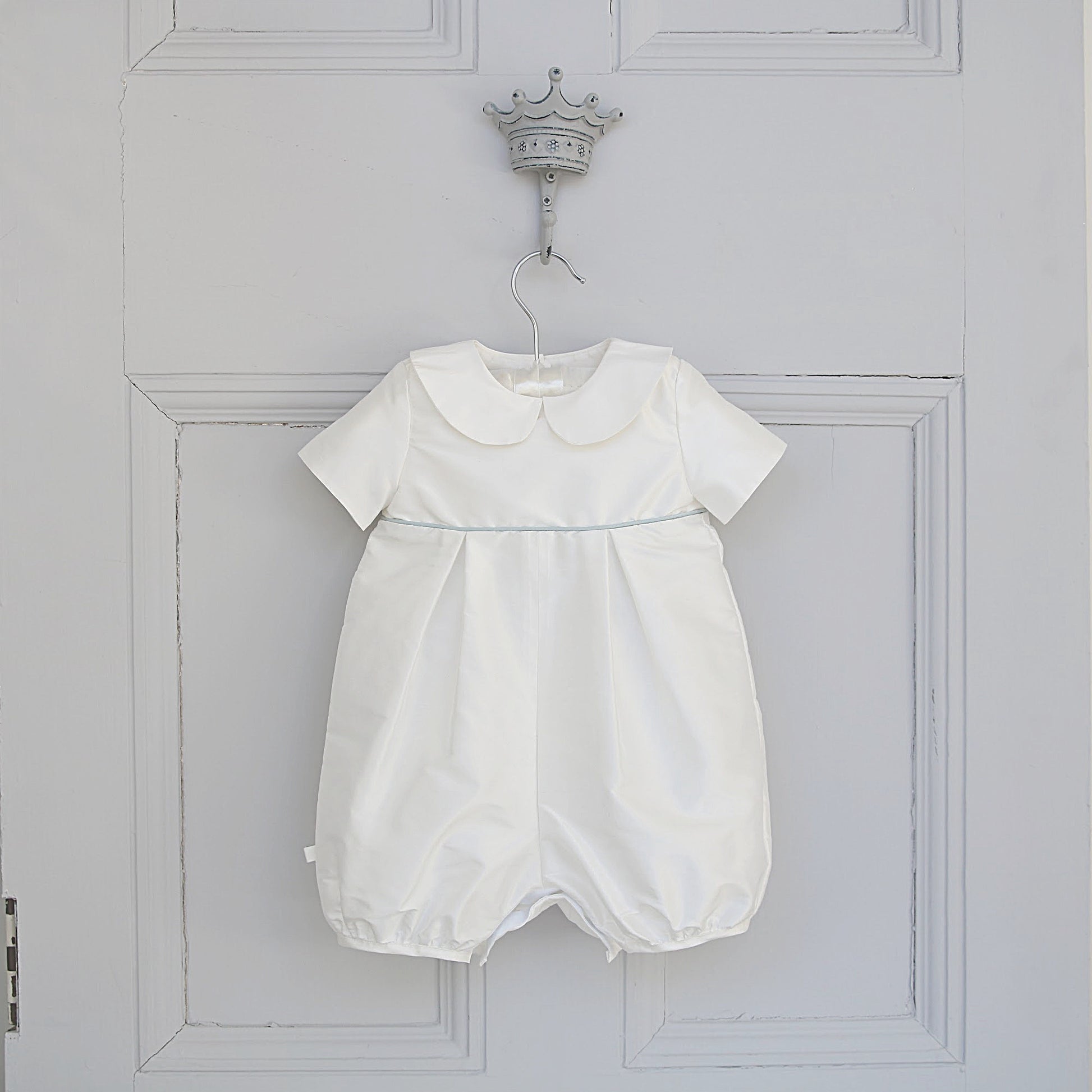 A white baby romper with short sleeves, a Peter Pan collar, and a subtle light blue piping detail at the waist, hanging on a decorative silver crown hook against a white paneled door.