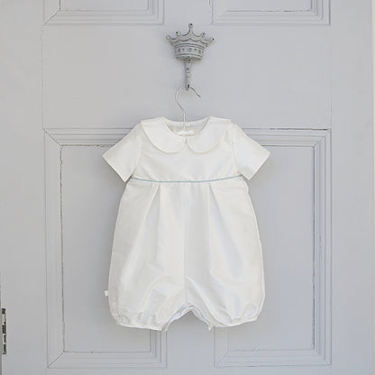 A white baby romper with short sleeves, a Peter Pan collar, and a subtle light blue piping detail at the waist, hanging on a decorative silver crown hook against a white paneled door.
