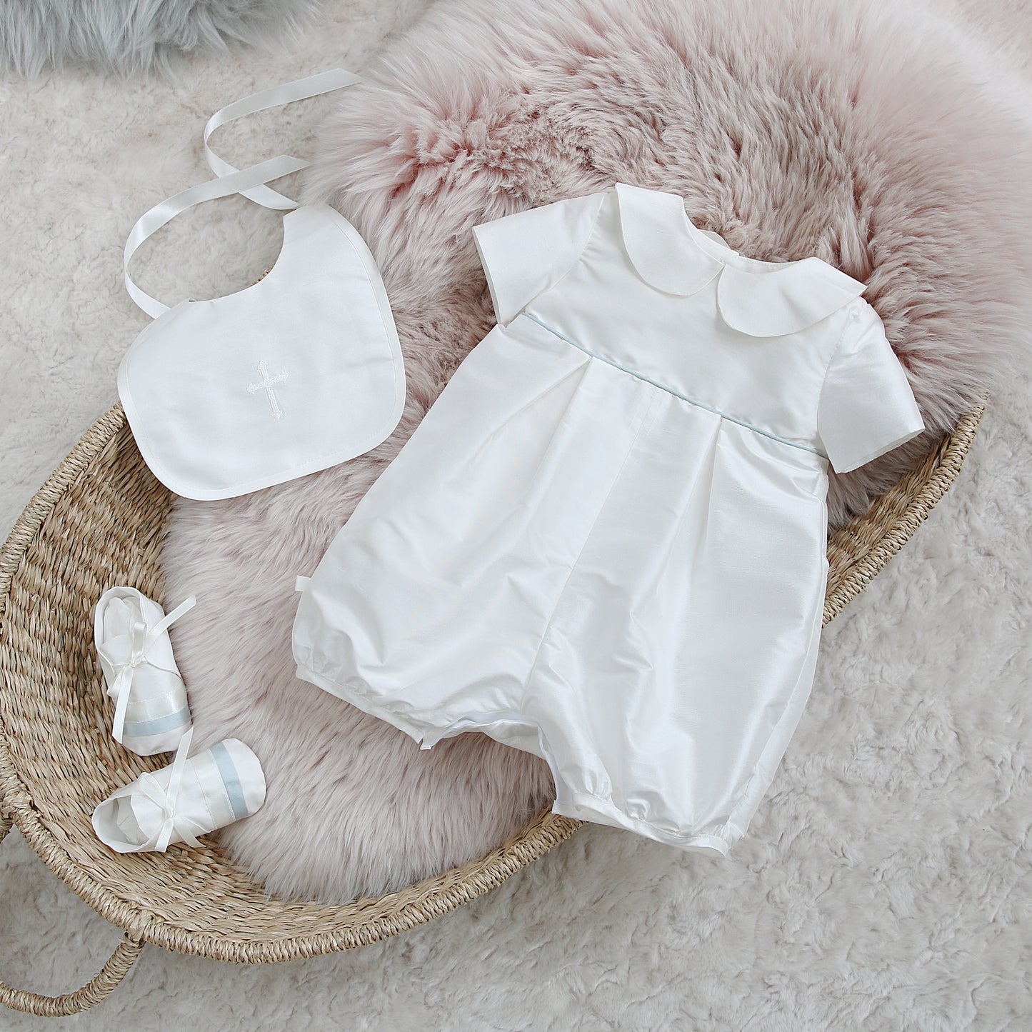 A baby first birthday outfit set including a white short-sleeve romper with a Peter Pan collar, a matching bib with a cross, and a pair of white booties, arranged on a pink faux fur cushion inside a wicker basket.