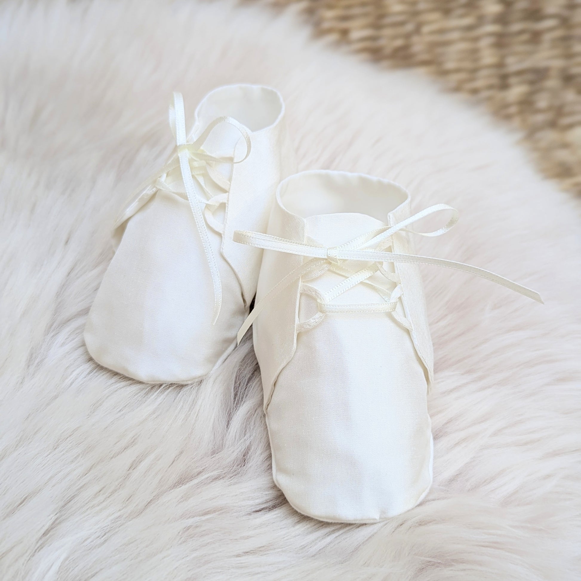 A pair of soft cream satin baby booties with ribbon laces, arranged on a fluffy white faux fur rug.