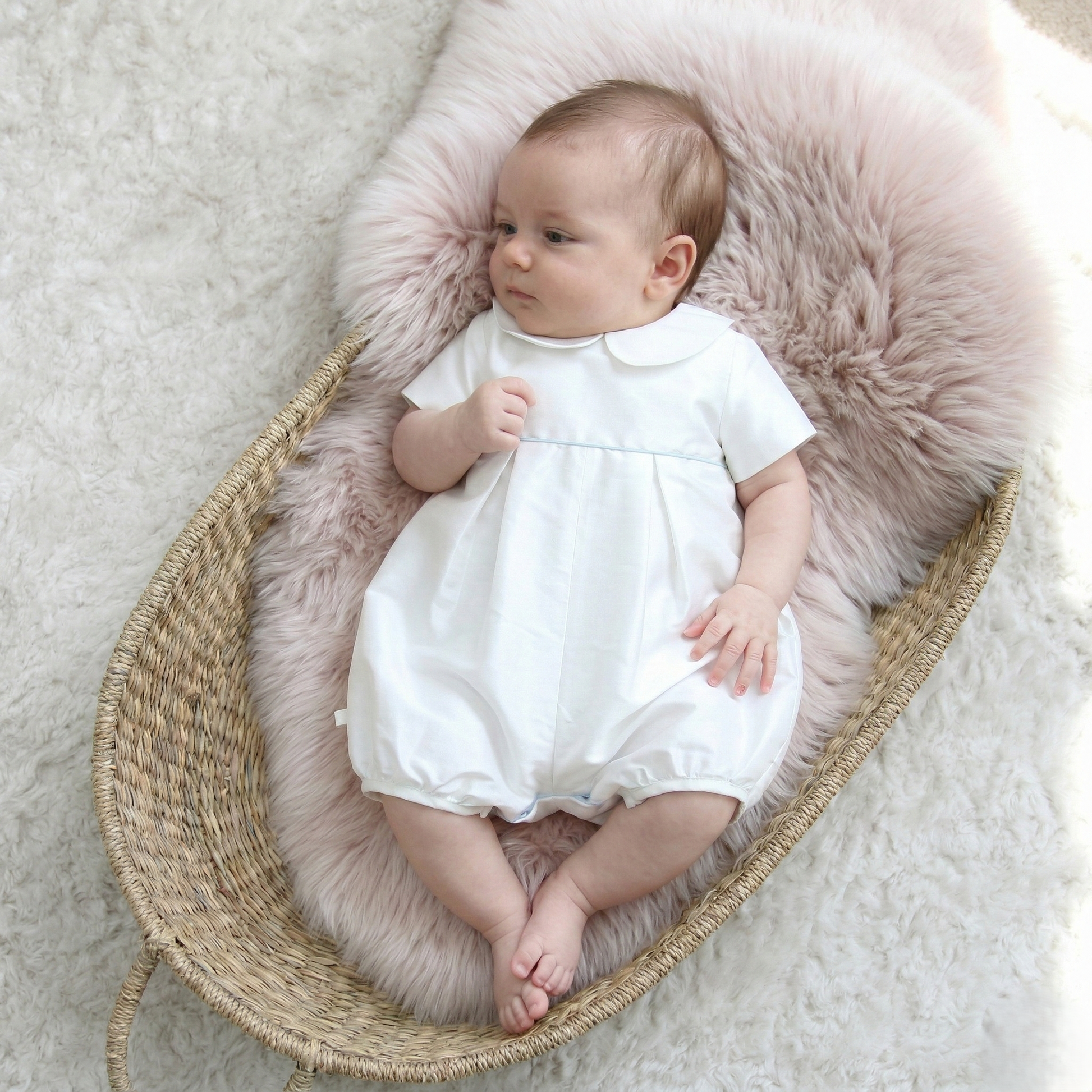 A baby wearing a white short-sleeve romper with a Peter Pan collar lies on a pink faux-fur cushion inside a woven rattan bassinet on a white rug.