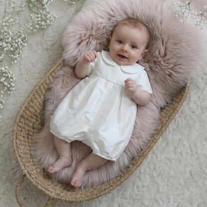 A baby wearing a white romper lies on a pink faux-fur cushion inside a woven basket, photographed from above on a soft cream rug with small white flowers around the edges.