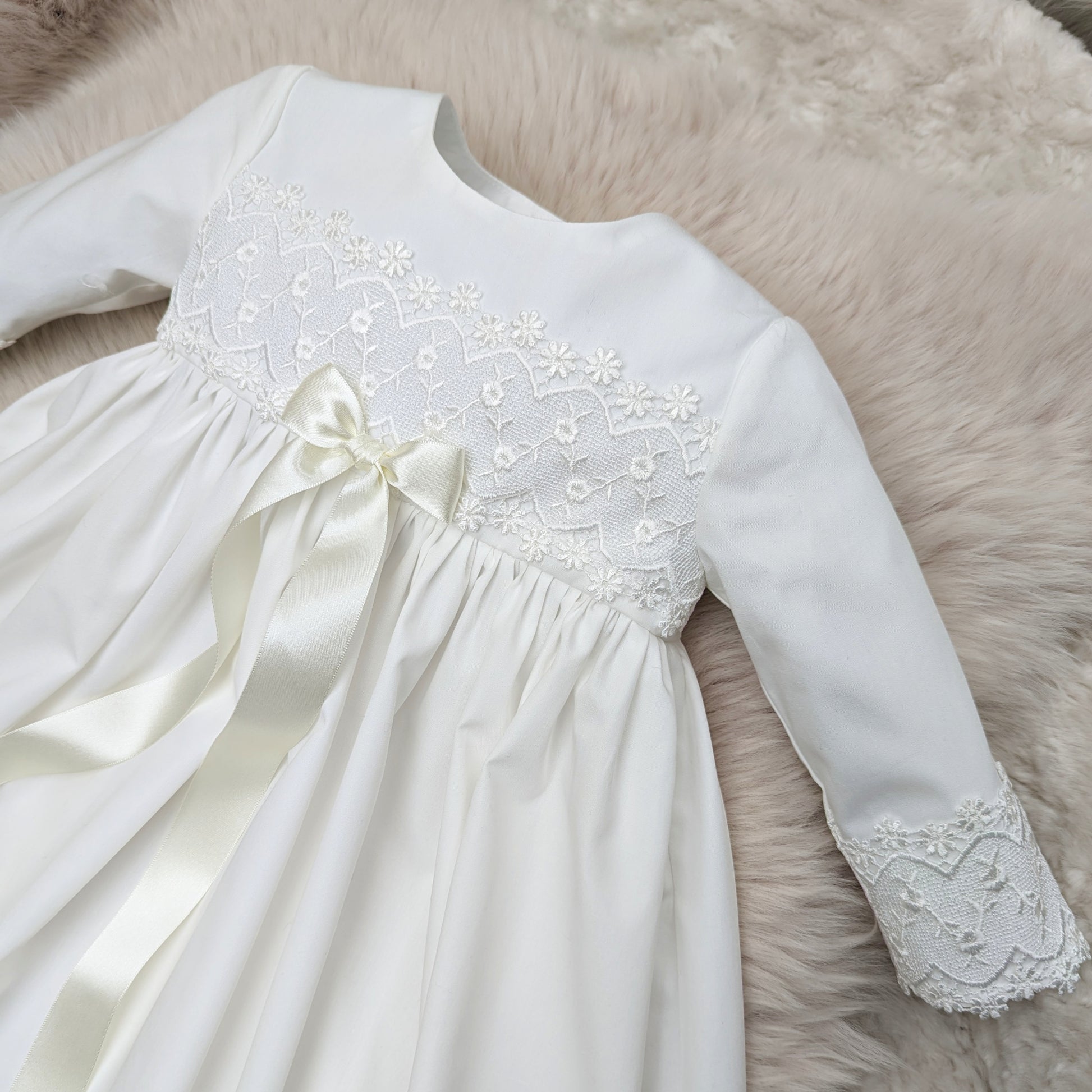 Close-up detail of the bodice on an ivory cotton Lana christening gown, showcasing the delicate lace embroidery and a long satin ribbon bow.