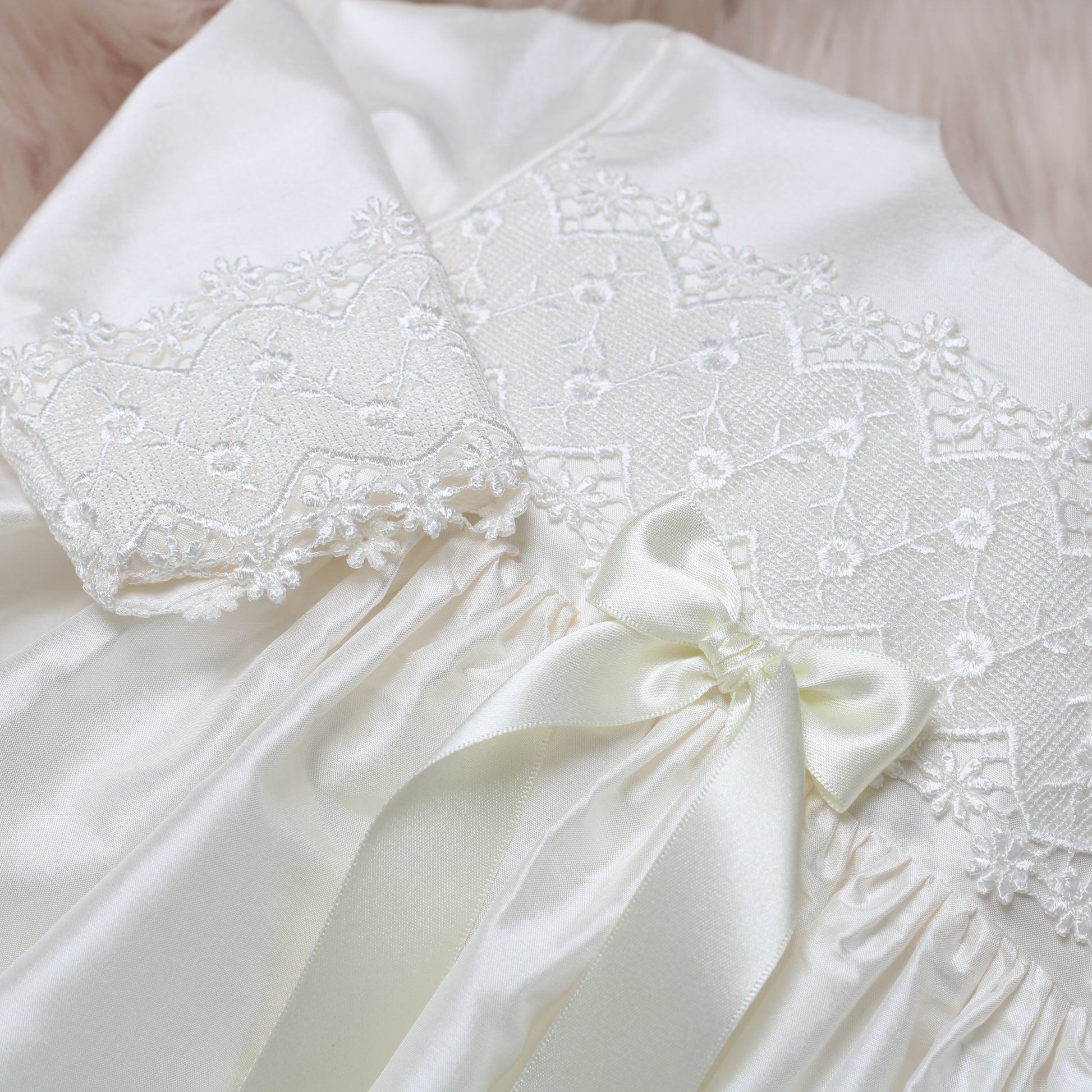 Close-up of the ivory silk Lara baby dress with delicate floral lace applique and a matching satin bow at the waist.