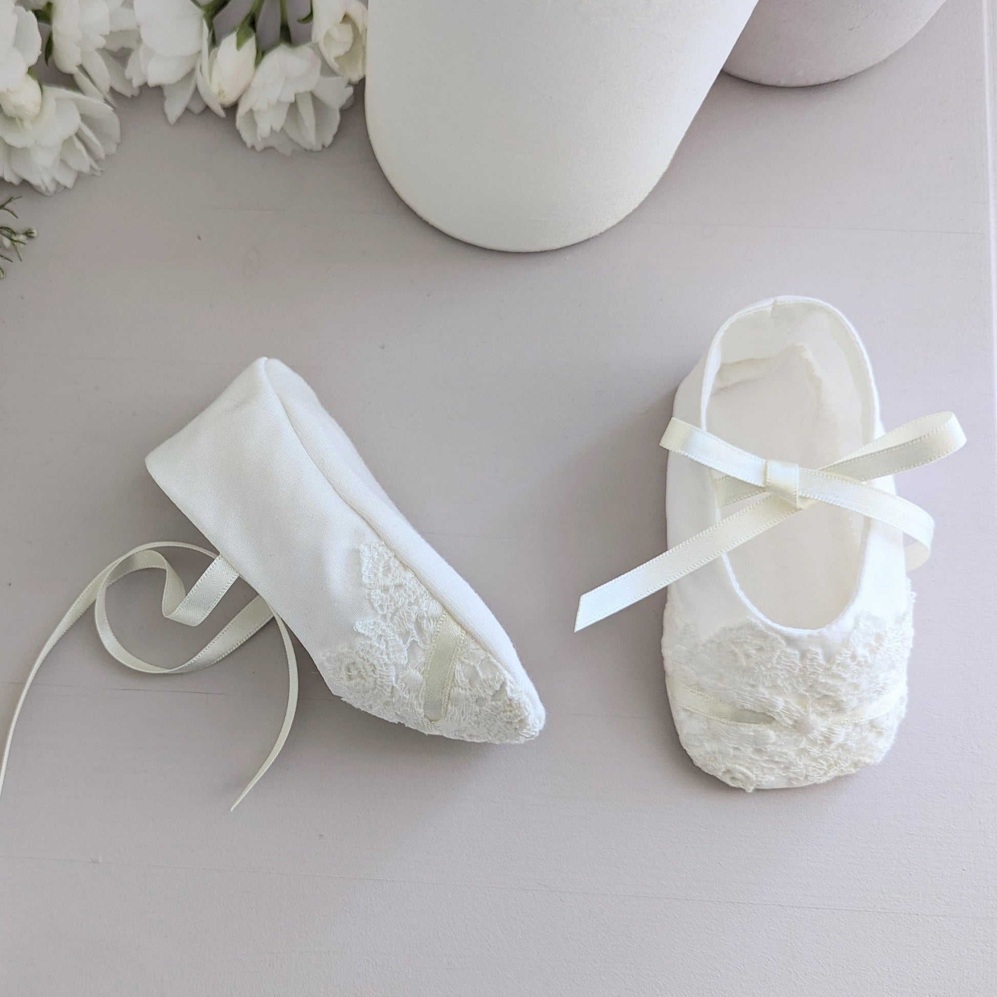 White baby shoes with ribbons on a light gray surface surrounded by white flowers.