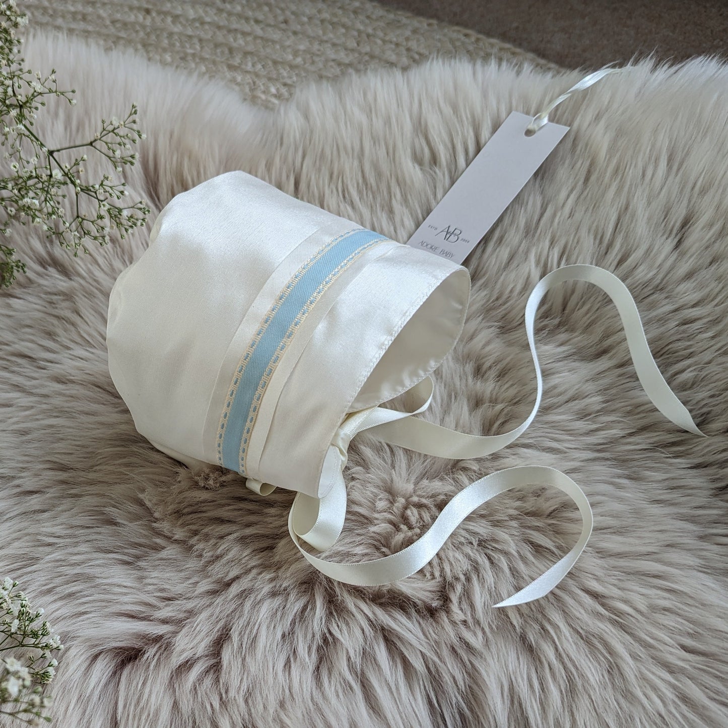 White baby bonnet with blue ribbon on a fluffy white surface with baby's breath flowers.