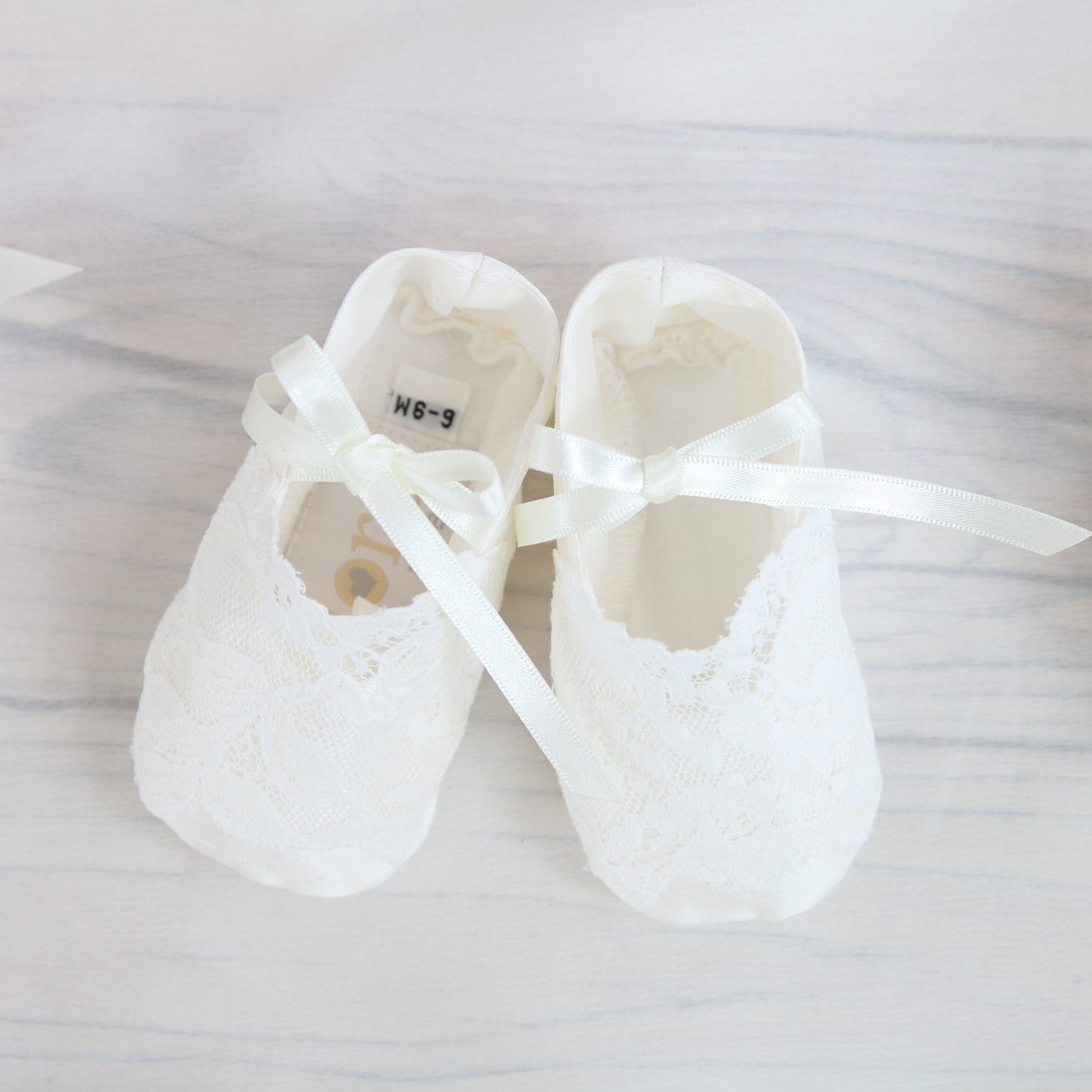 Serenity cotton christening booties with lace trim, displayed on a rustic white wooden floor background